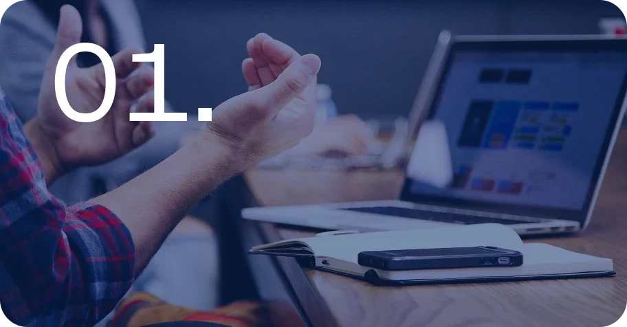Hands in front of a desk with a laptop, a notebook and a smartphone with the number 01. displayed in the foreground.